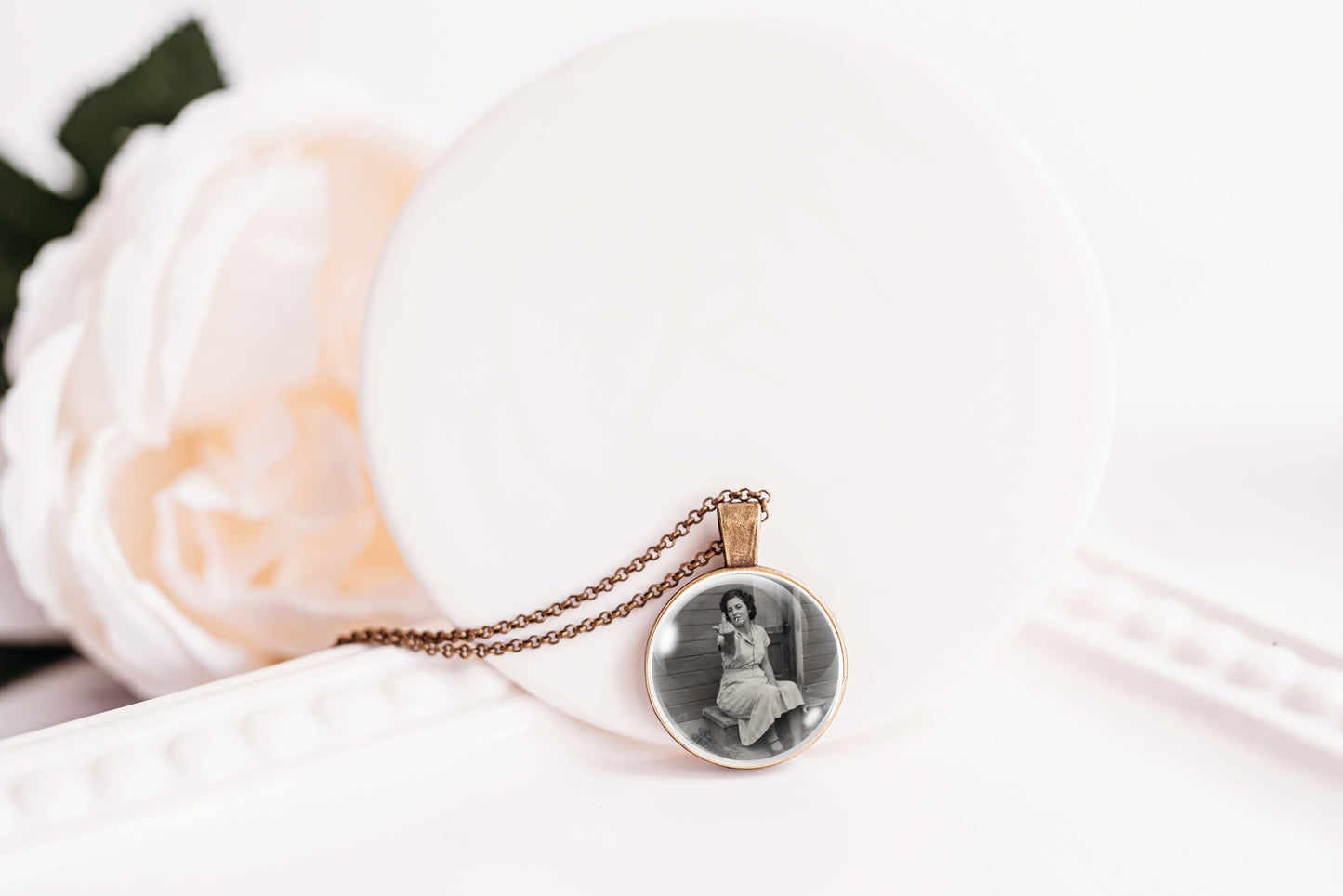 A round pendant necklace on a delicate chain, featuring a black and white portrait photo of a woman, displayed on a white circular stand with a soft white rose in the background.