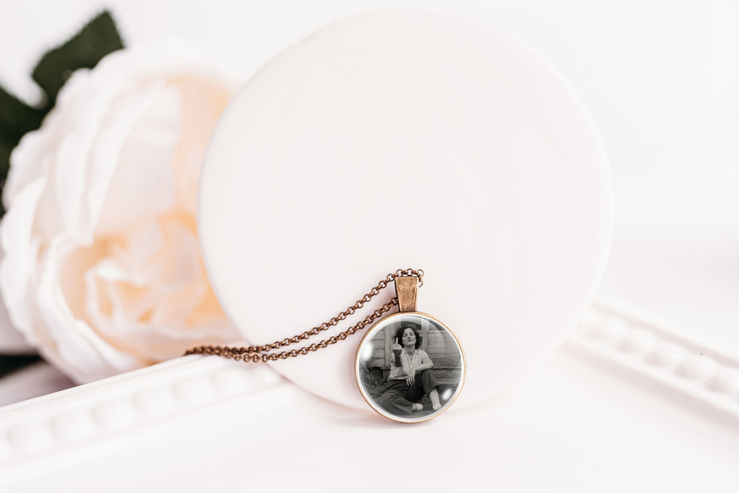 A round pendant necklace with a black-and-white photo of a woman, hanging from a bronze-toned chain, displayed on a white circular stand next to a soft white rose.