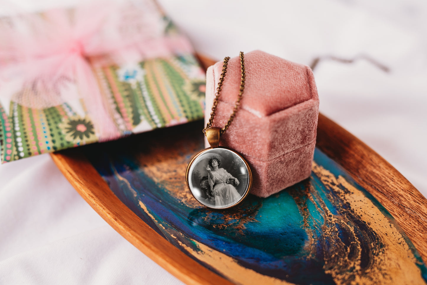 A round pendant necklace with a black-and-white portrait photo in a silver-toned bezel, hanging from a gold-tone chain and resting on a pink velvet jewelry box. The box sits on a decorative wooden tray with blue and gold marbling, with a patterned fabric bag in the background.