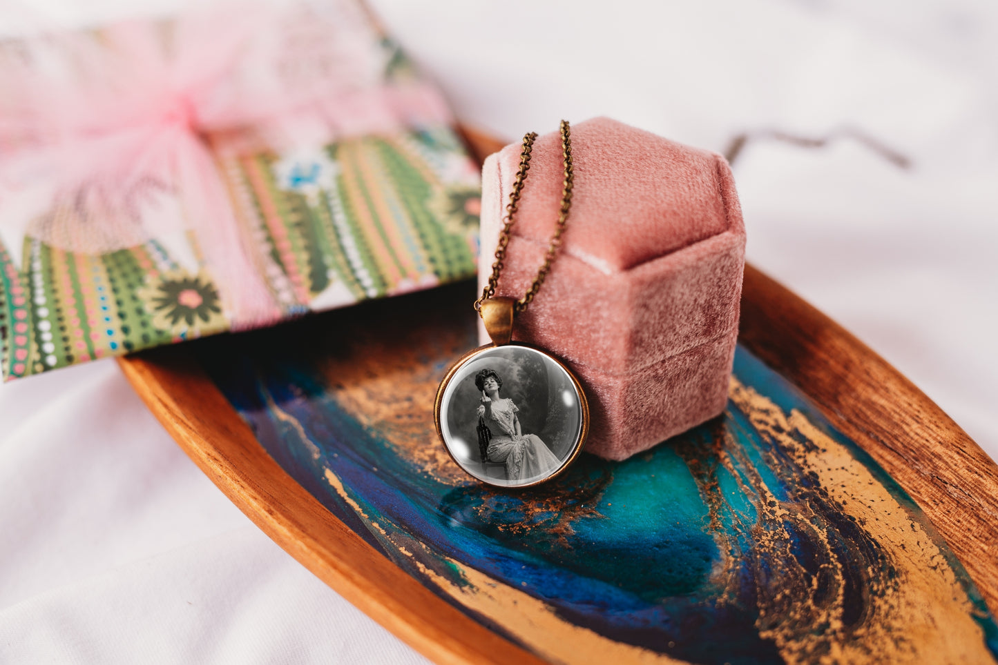 A custom round pendant necklace with a black-and-white portrait photo, displayed on a pink velvet ring box resting on a decorative wooden tray.
