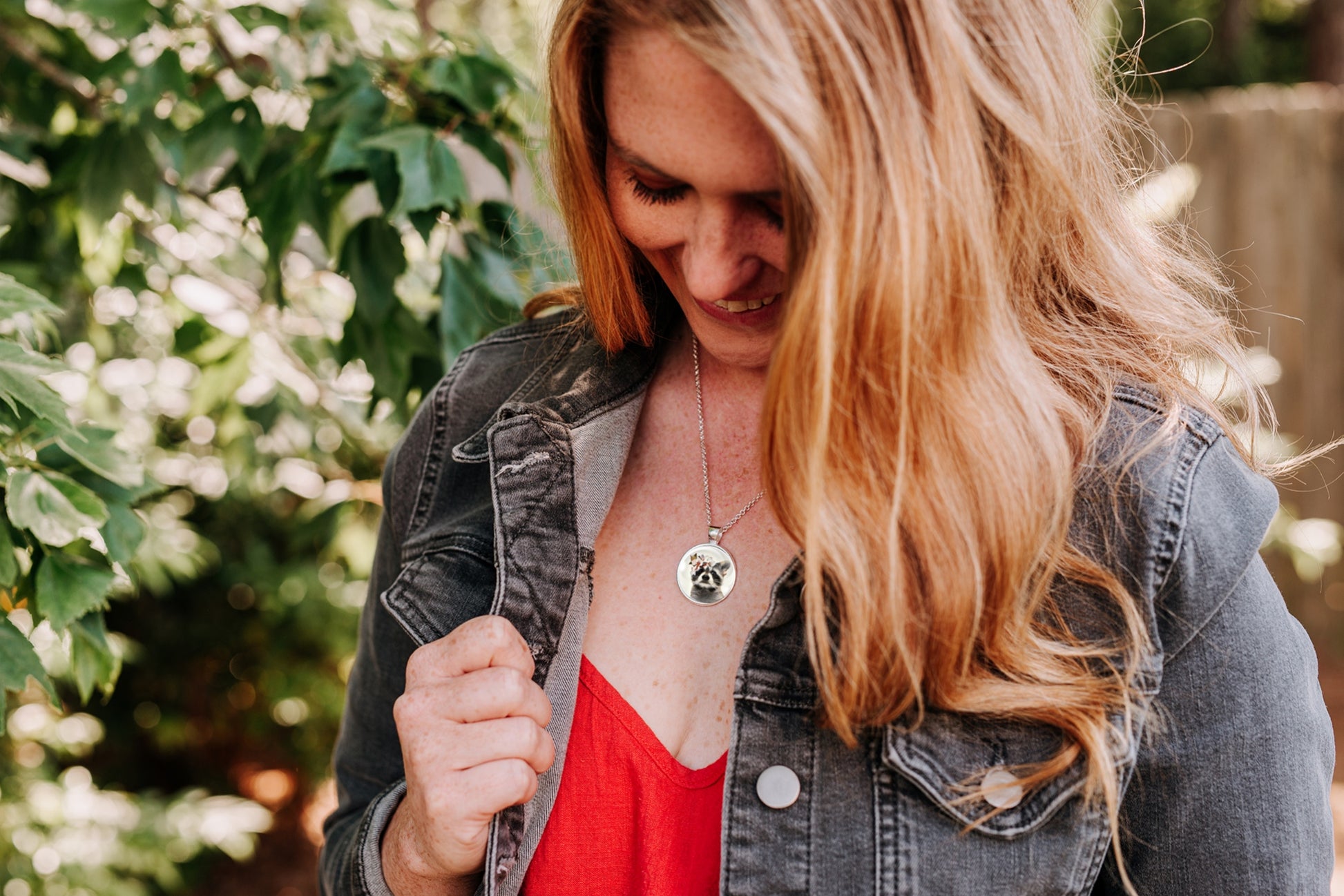 Necklace with a pendant featuring a raccoon wearing a flower crown shown worn by a woman wearing a red top and black denim jacket