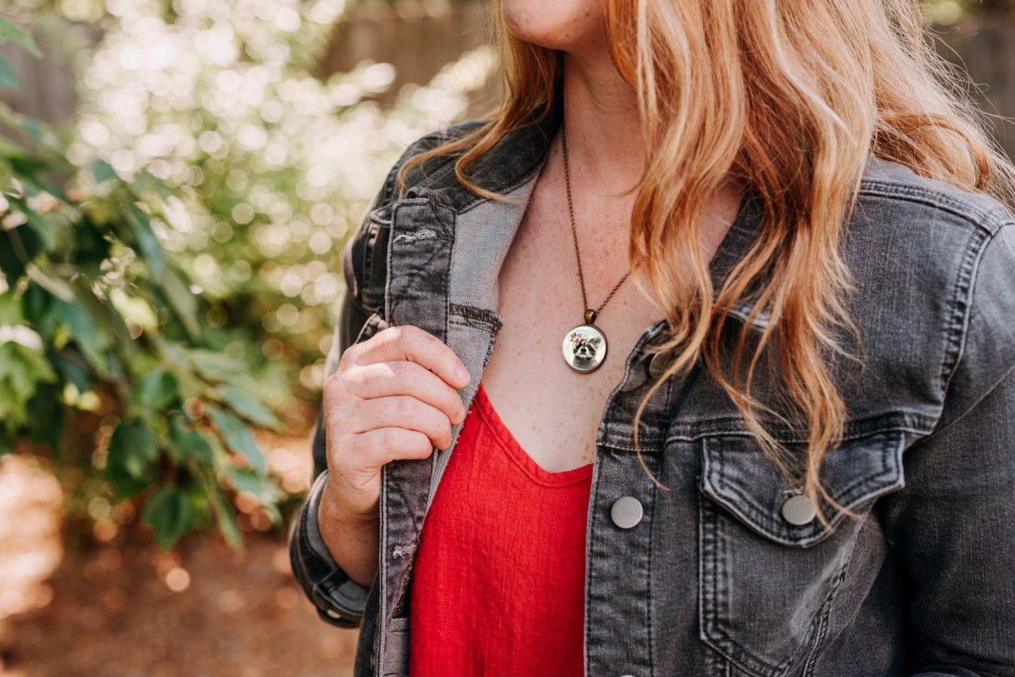 Necklace with a pendant featuring a raccoon wearing a flower crown shown worn by a woman wearing a red top and black denim jacket