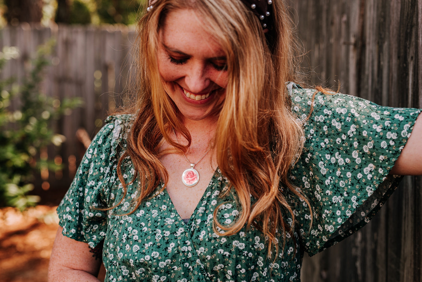 Woman wearing a pink rose pendant necklace and green floral dress next to a fence