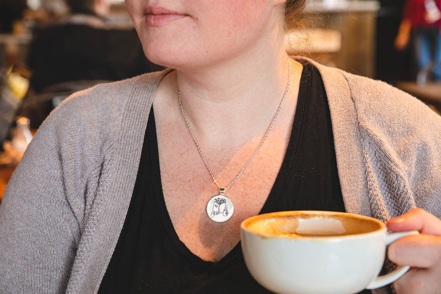 Woman wearing a pendant necklace with a pendant featuring an image of a hand holding a book with branches coming out of the top and a cup, holding a cup of coffee with a blurred background