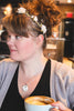 Woman with a floral headband wearing a bookish pendant necklace and holding a mug in an indoor setting