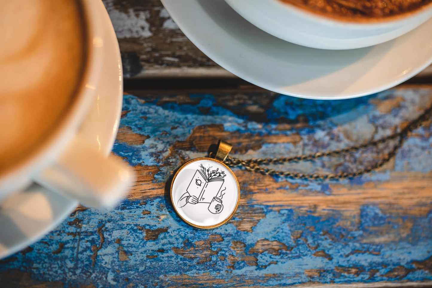 Necklace with a pendant featuring an image of a hand holding a book with branches coming out of the top and a cup, resting on a textured wooden surface with two coffees next to it.