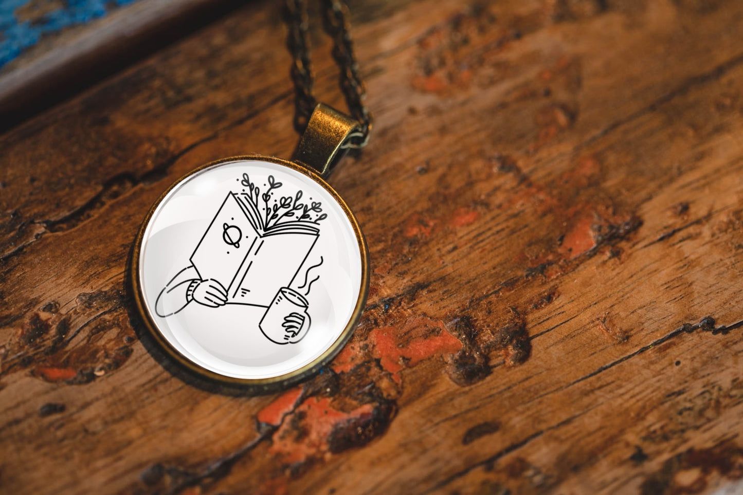 Necklace with a pendant featuring an image of a hand holding a book with branches coming out of the top and a cup, resting on a textured wooden surface.