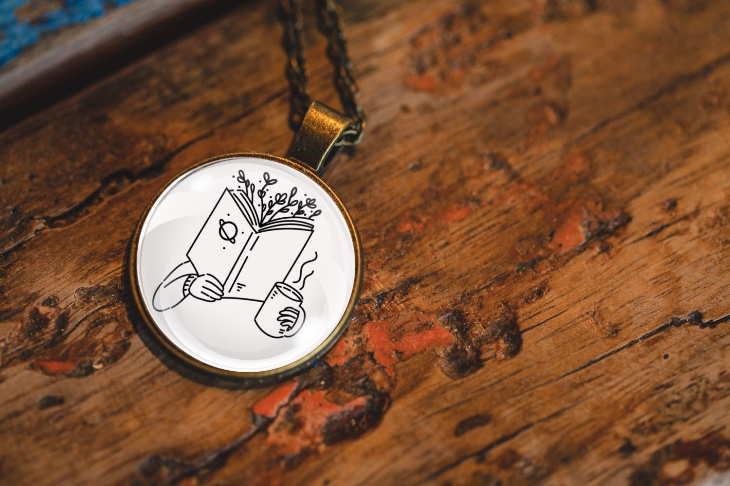 Necklace with a pendant featuring an image of a hand holding a book with branches coming out of the top and a cup, resting on a textured wooden surface.