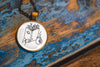 Necklace with a pendant featuring an image of a hand holding a book with branches coming out of the top and a cup, resting on a textured wooden surface.