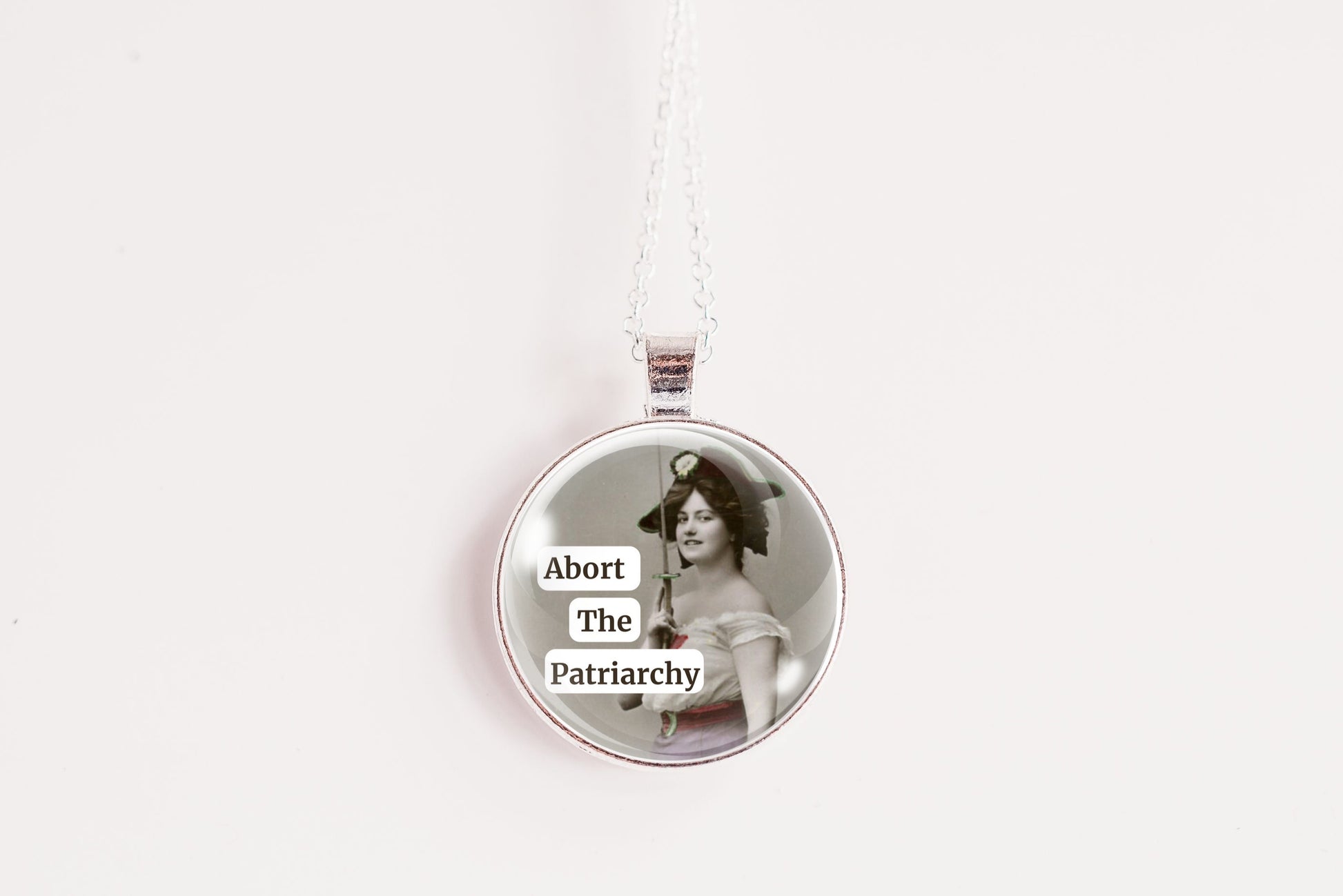 Product photo of a 1-inch glass cabochon pendant necklace featuring “Abort the Patriarchy” with a vintage-style woman holding a sword on a 20-inch chain.