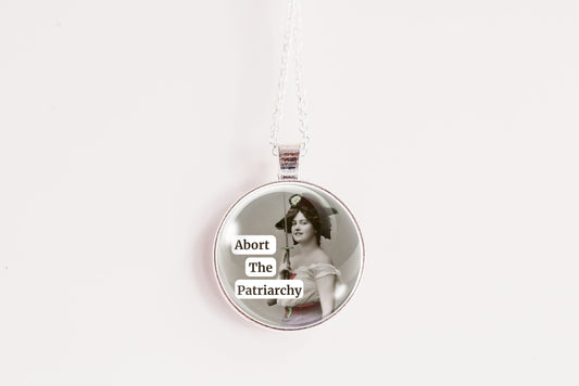 Product photo of a 1-inch glass cabochon pendant necklace featuring “Abort the Patriarchy” with a vintage-style woman holding a sword on a 20-inch chain.