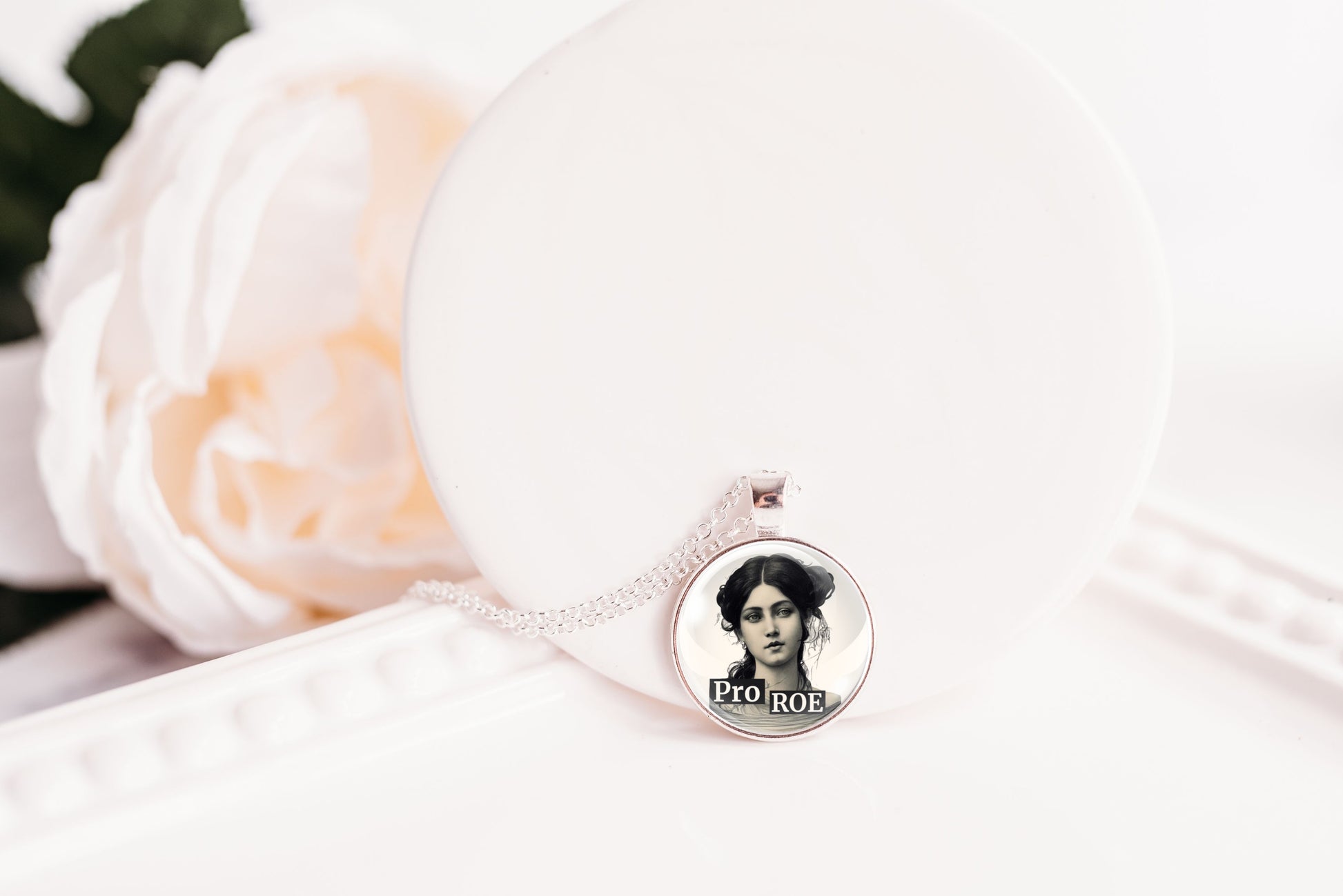 A silver-toned necklace with a round pendant displaying a black-and-white portrait photo, resting on a white display stand next to a soft white rose.