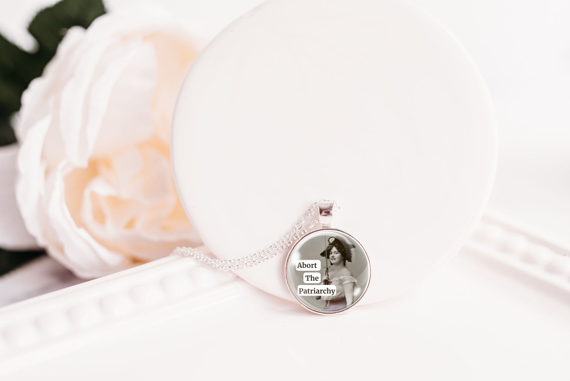Product photo of a 1-inch glass cabochon pendant necklace featuring “Abort the Patriarchy” with a vintage-style woman holding a sword on a 20-inch chain.