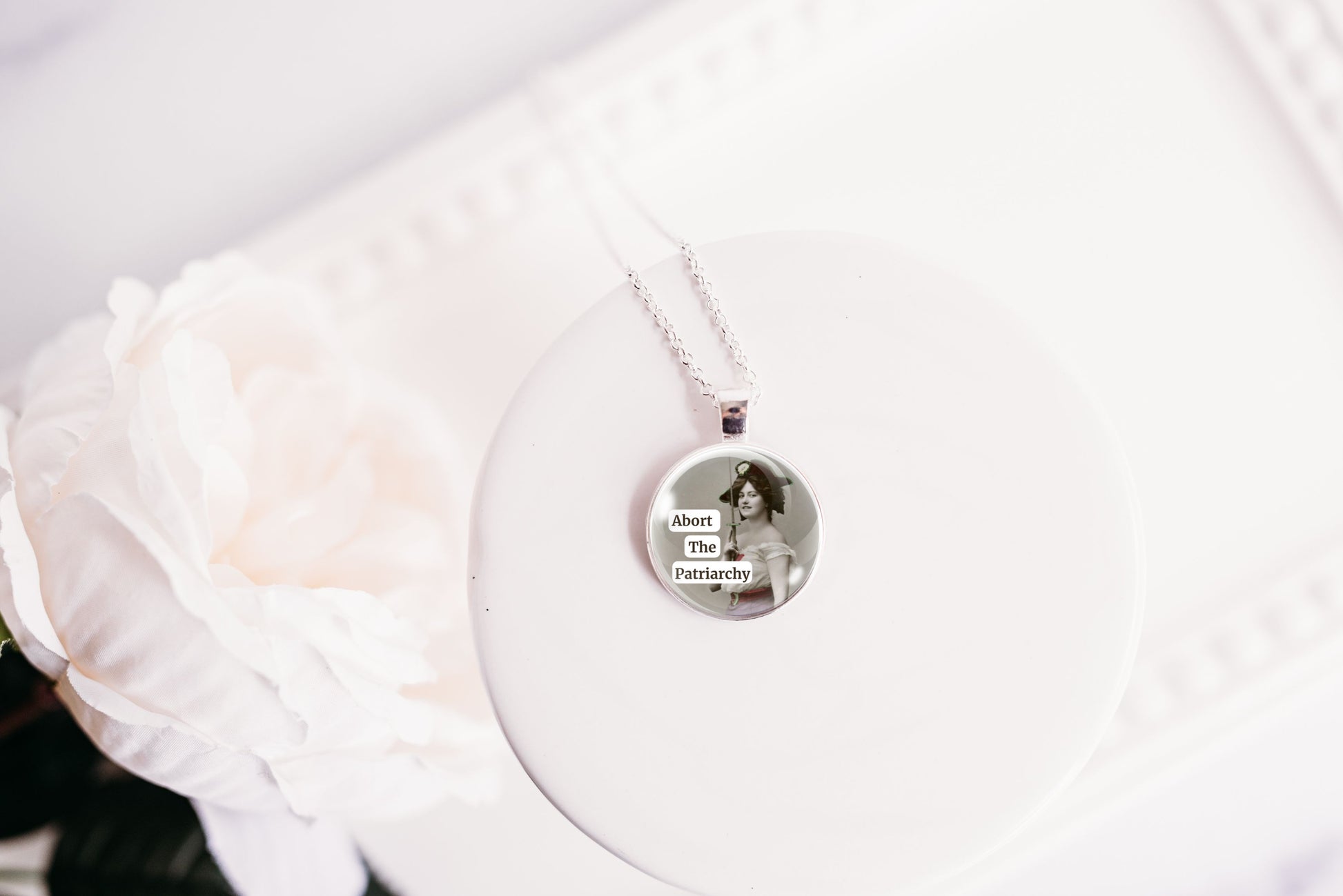 Product photo of a 1-inch glass cabochon pendant necklace featuring “Abort the Patriarchy” with a vintage-style woman holding a sword on a 20-inch chain.