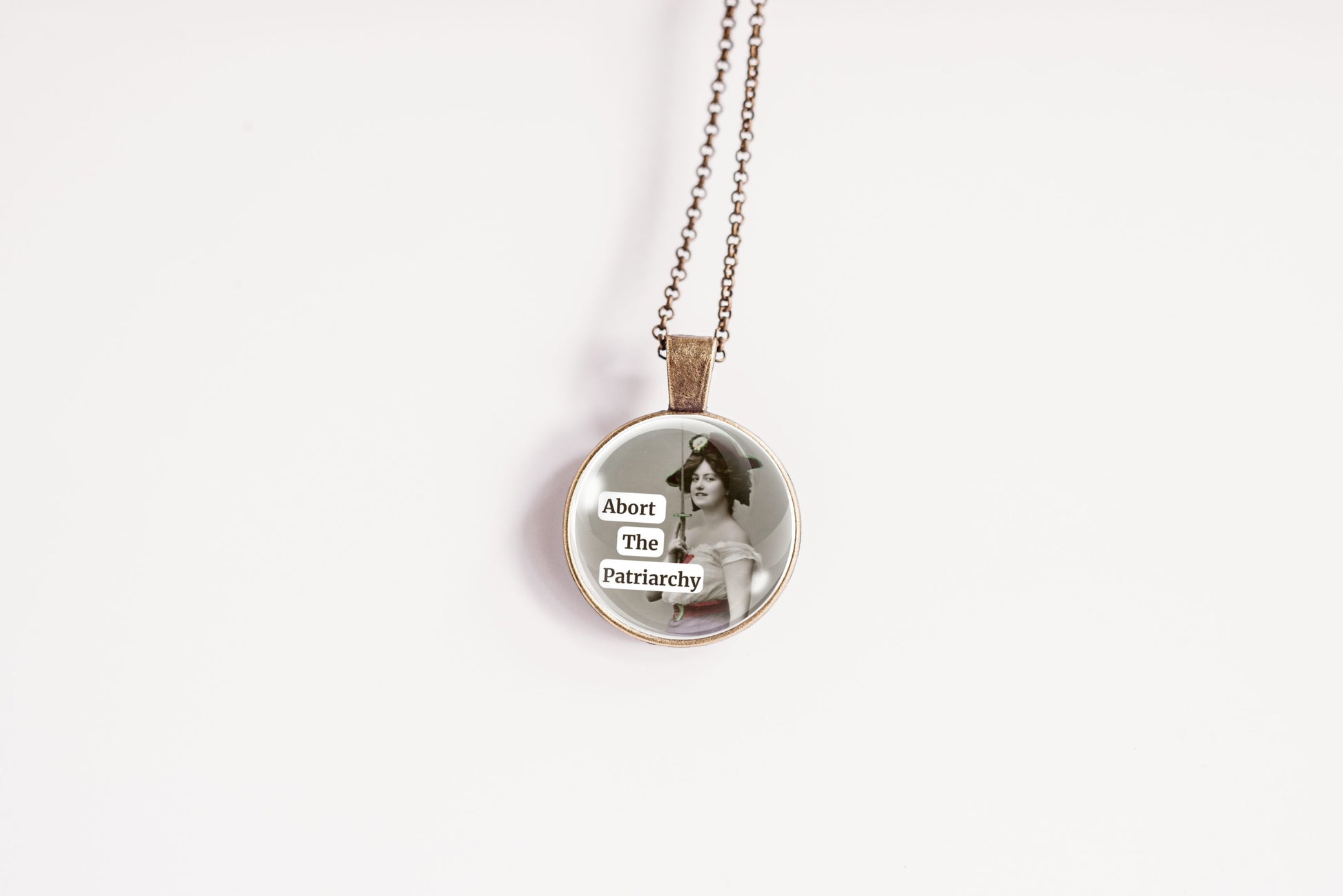 Product photo of a 1-inch glass cabochon pendant necklace featuring “Abort the Patriarchy” with a vintage-style woman holding a sword on a 20-inch chain.