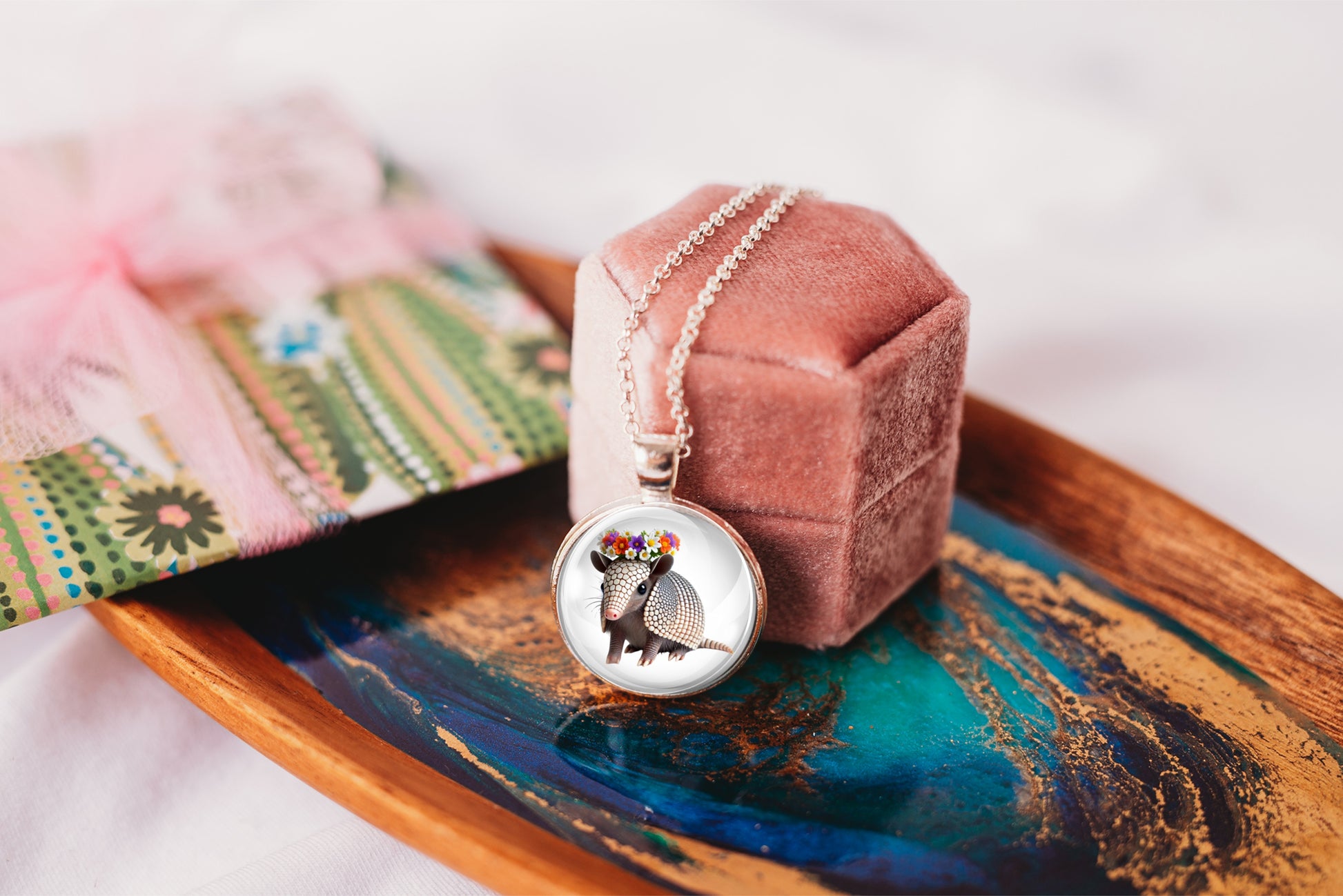 Necklace with a pendant featuring an armadillo wearing a flower crown shown against a pink velvet jewelry box on a decorative tray