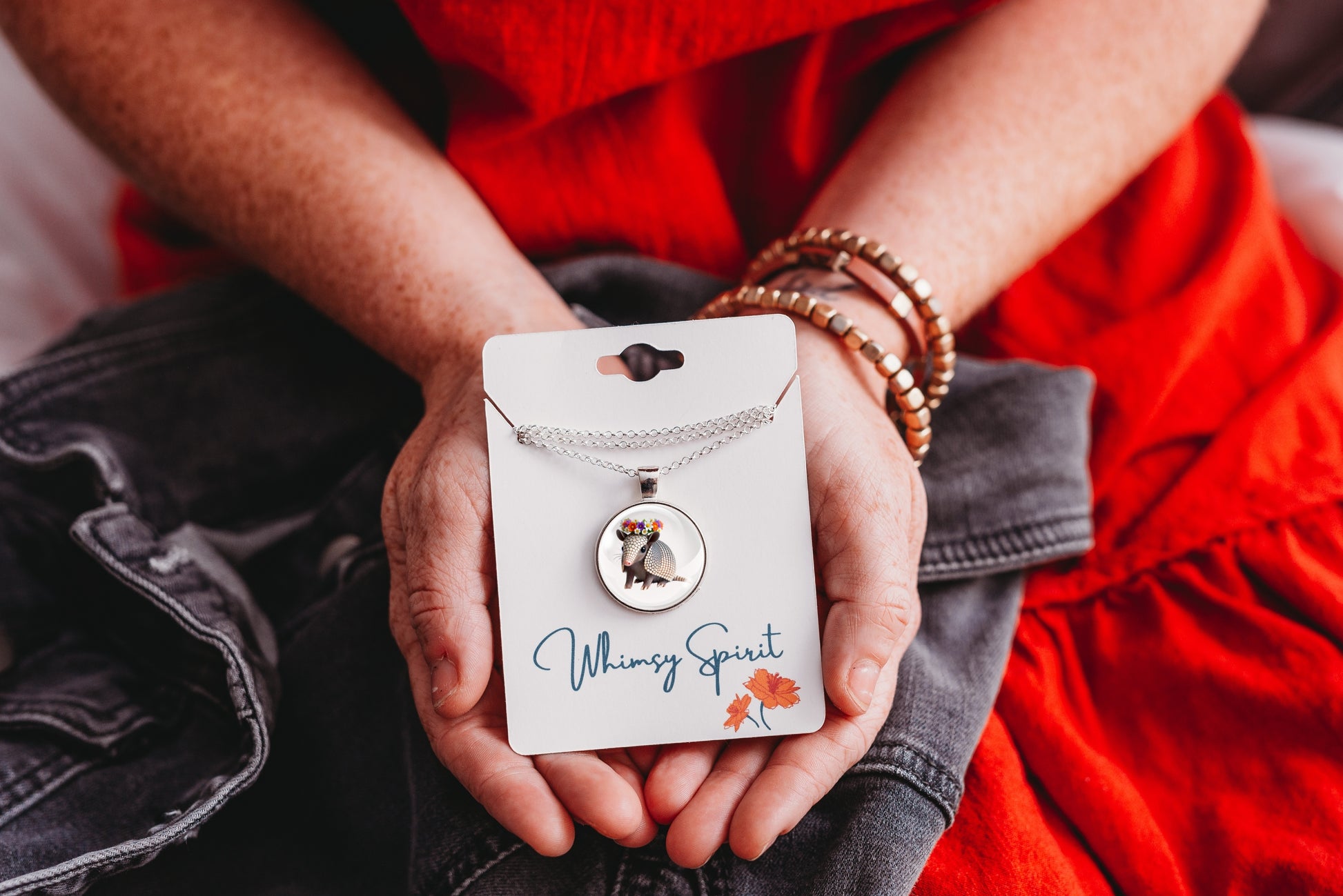 Person holding an armadillo necklace with a floral pendant on a branded card, wearing a red garment and black pants.