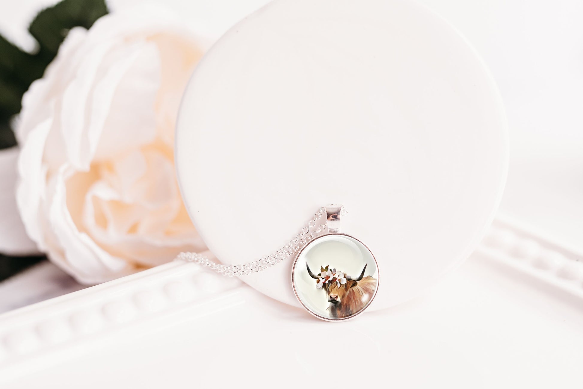 Necklace with a pendant featuring a highland cow design, displayed on a white surface with flowers.