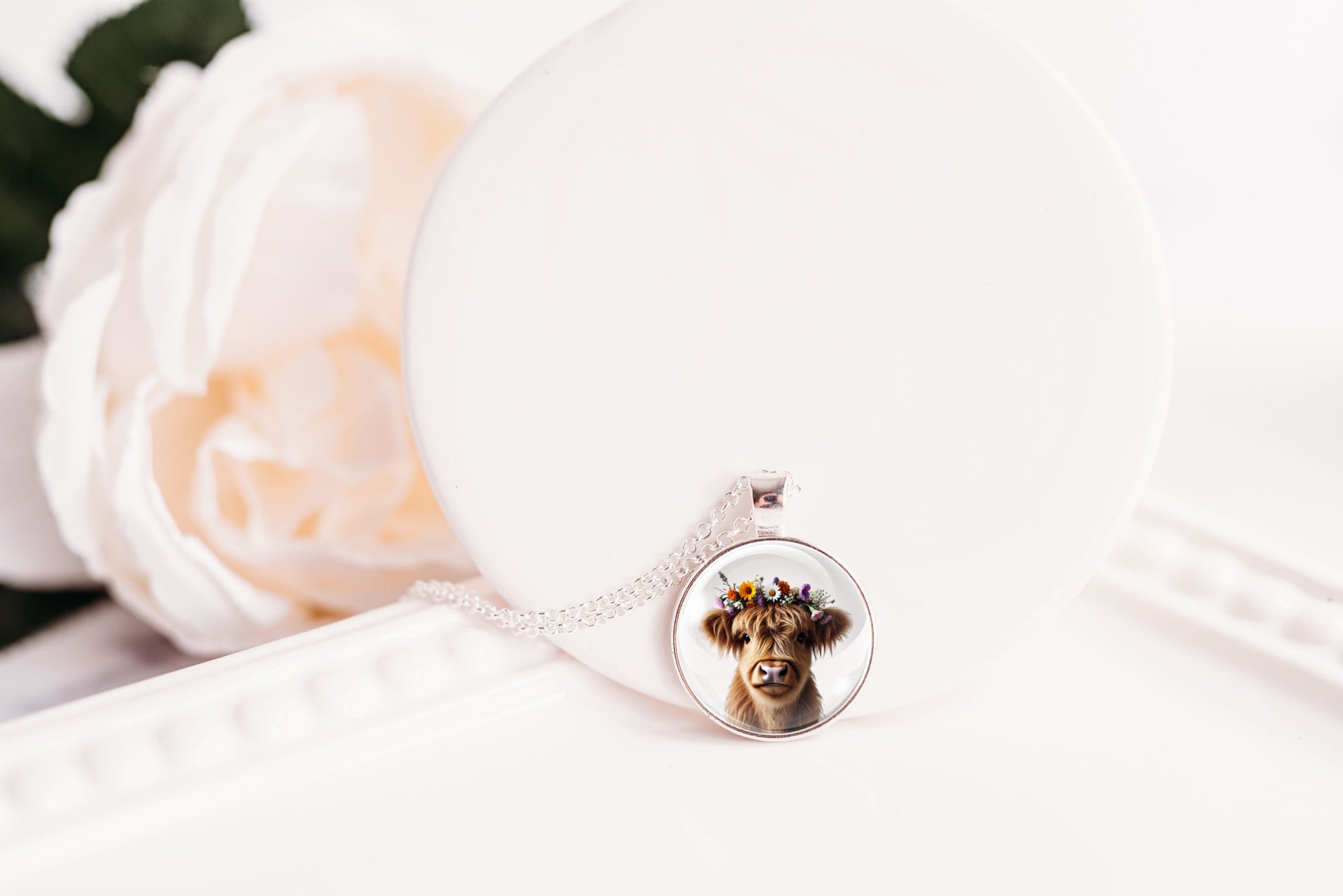 Necklace with a pendant featuring a highland cow with a flower crown on a white surface with a flower in the background