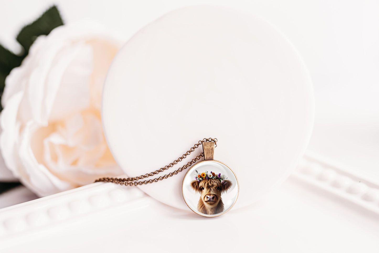 Necklace with a pendant featuring a highland cow with a flower crown on a white surface with a flower in the background
