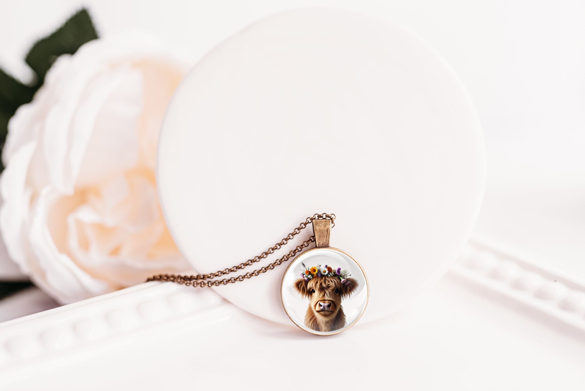 Necklace with a pendant featuring a highland cow with a flower crown on a white surface with a flower in the background