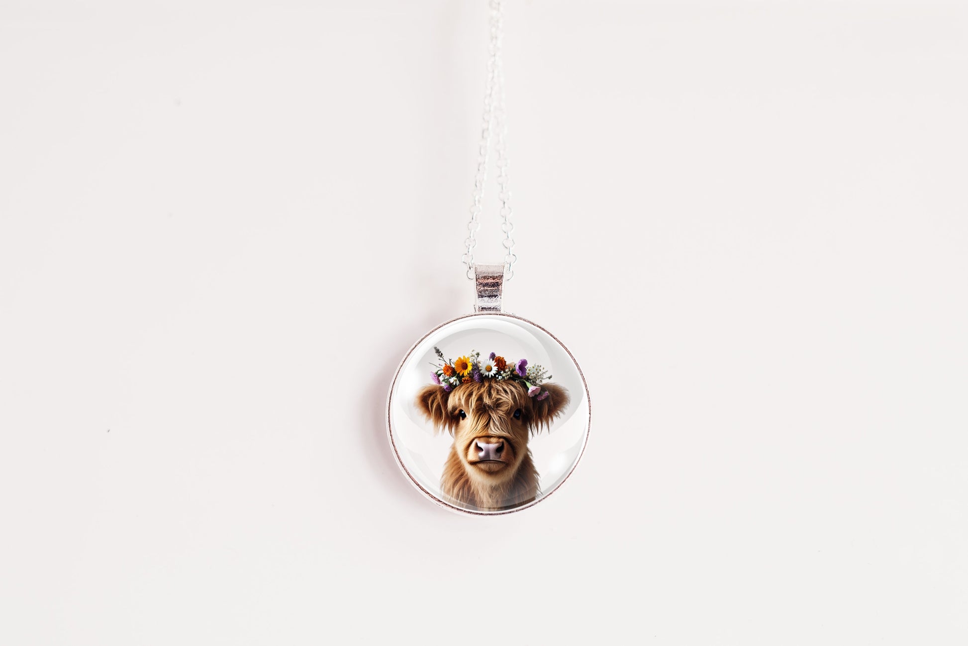 Necklace with a pendant featuring a highland cow with a flower crown on a white surface with a flower in the background