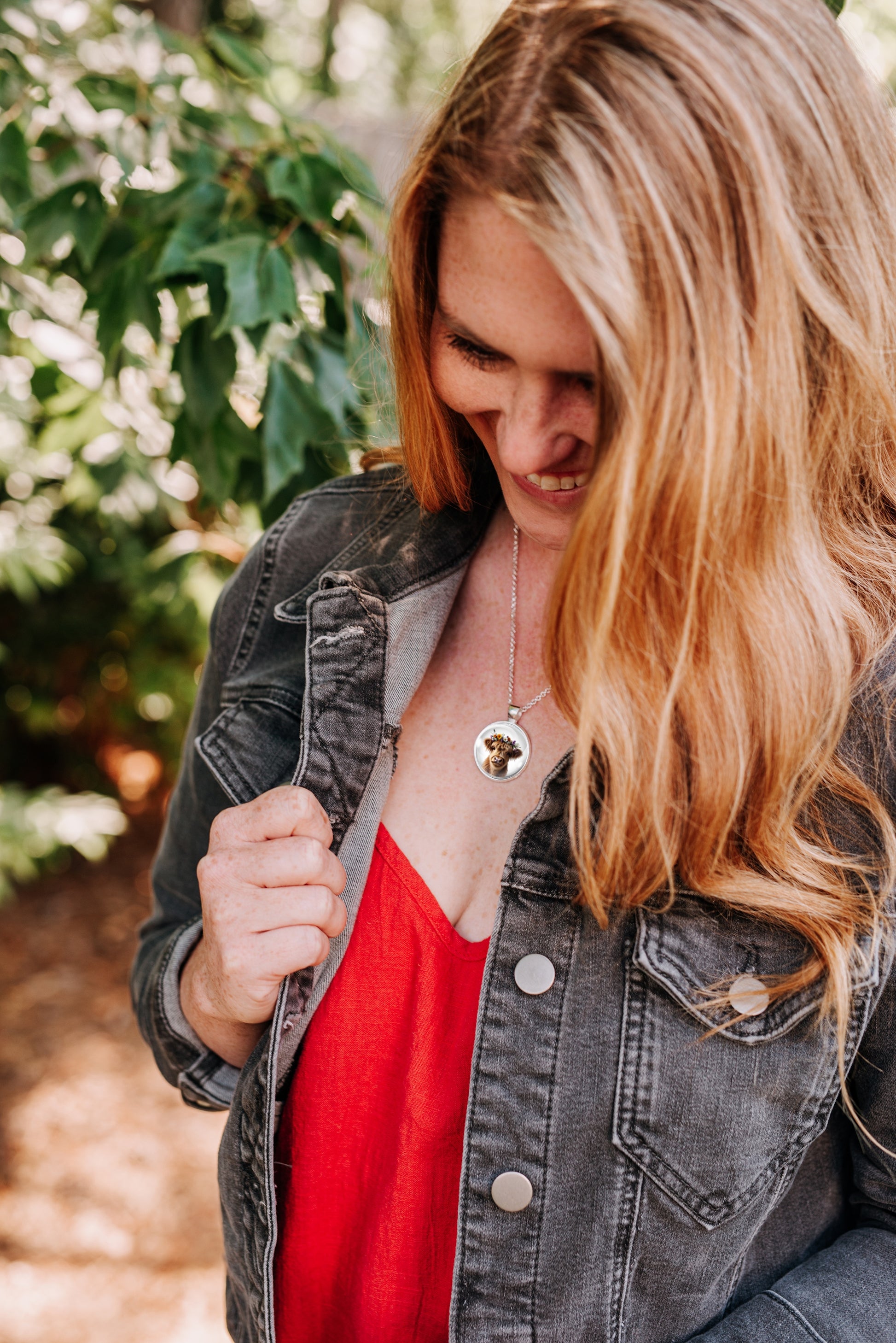 Woman in a red top and a black denim jacket wearing a highland cow pendant necklace standing outdoors next to a wooden fence.