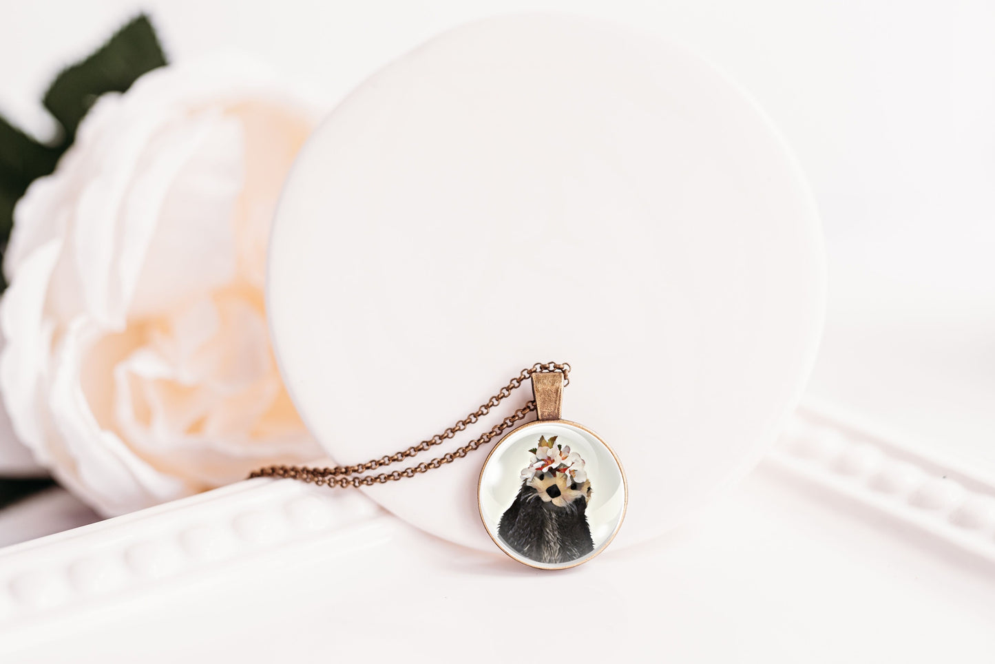 Necklace with a pendant featuring an otter wearing a flower crown design on a white surface with a blurred flower in the background