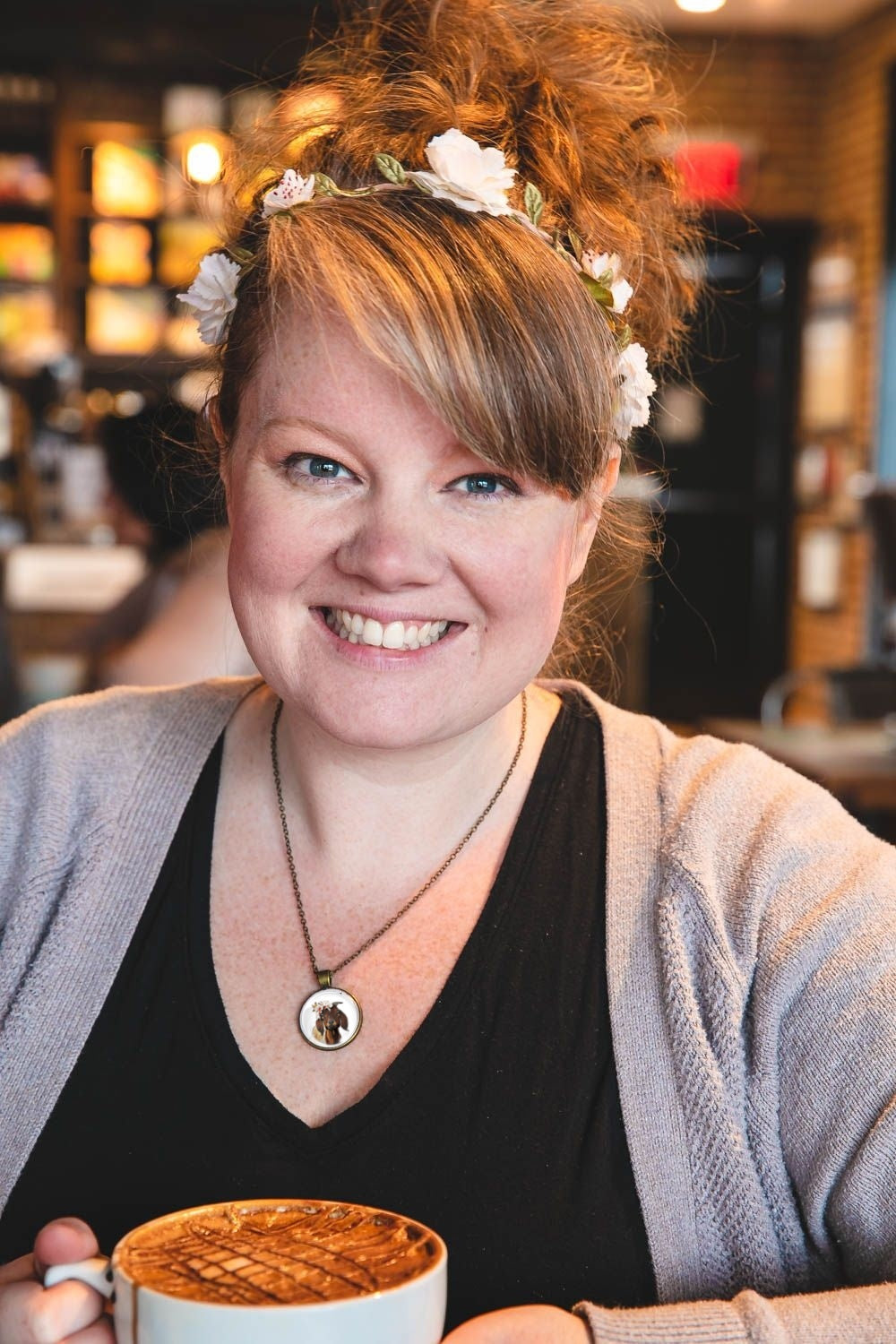 Woman with a floral headband wearing a flower crown goat pendant necklace holding a mug in a casual setting