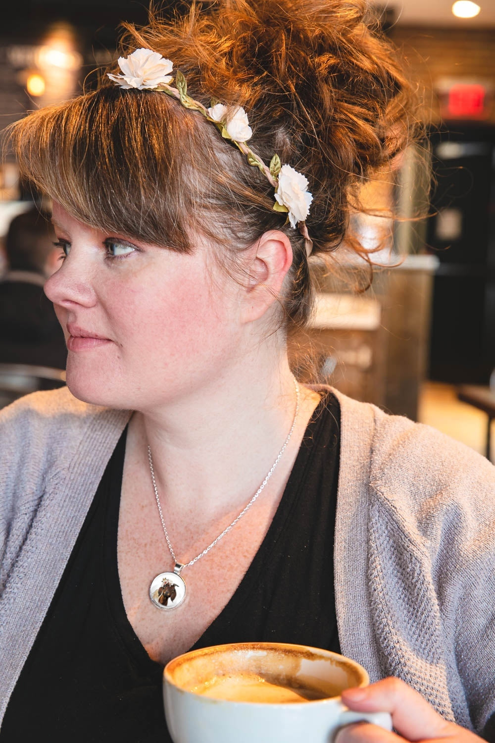 Woman with a floral headband wearing a flower crown goat pendant necklace holding a mug in a casual setting