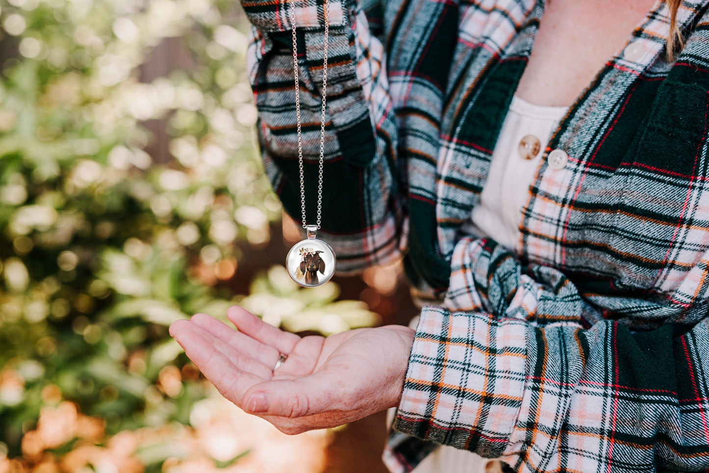 Woman wearing a plaid shirt holding a necklace with a goat pendant outdoors.