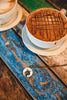 Cappuccino with a lattice pattern on top served in a white cup on a blue wooden surface next to a pendant necklace featuring a goat wearing a flower crown.