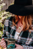 Woman wearing a black hat and plaid shirt, and a pendant necklace featuring an image of a fox wearing a flower crown, holding a mug outdoors.