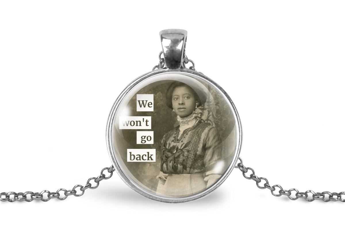 A silver pendant necklace featuring a circular glass cabochon with a sepia-toned vintage portrait of a woman and the text 'We won't go back' in white lettering, shown on a white background with a silver chain.