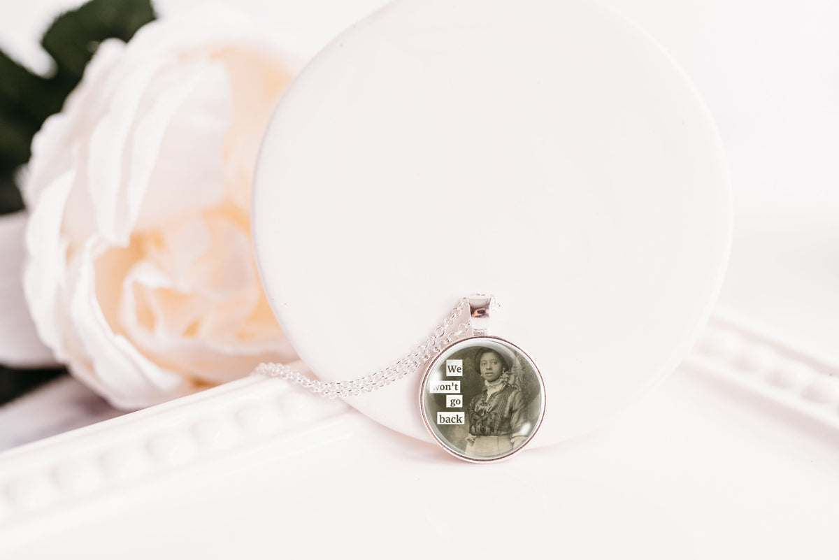 A silver-toned necklace with a round pendant displaying a photo of a woman of color, resting on a white display stand next to a white rose.