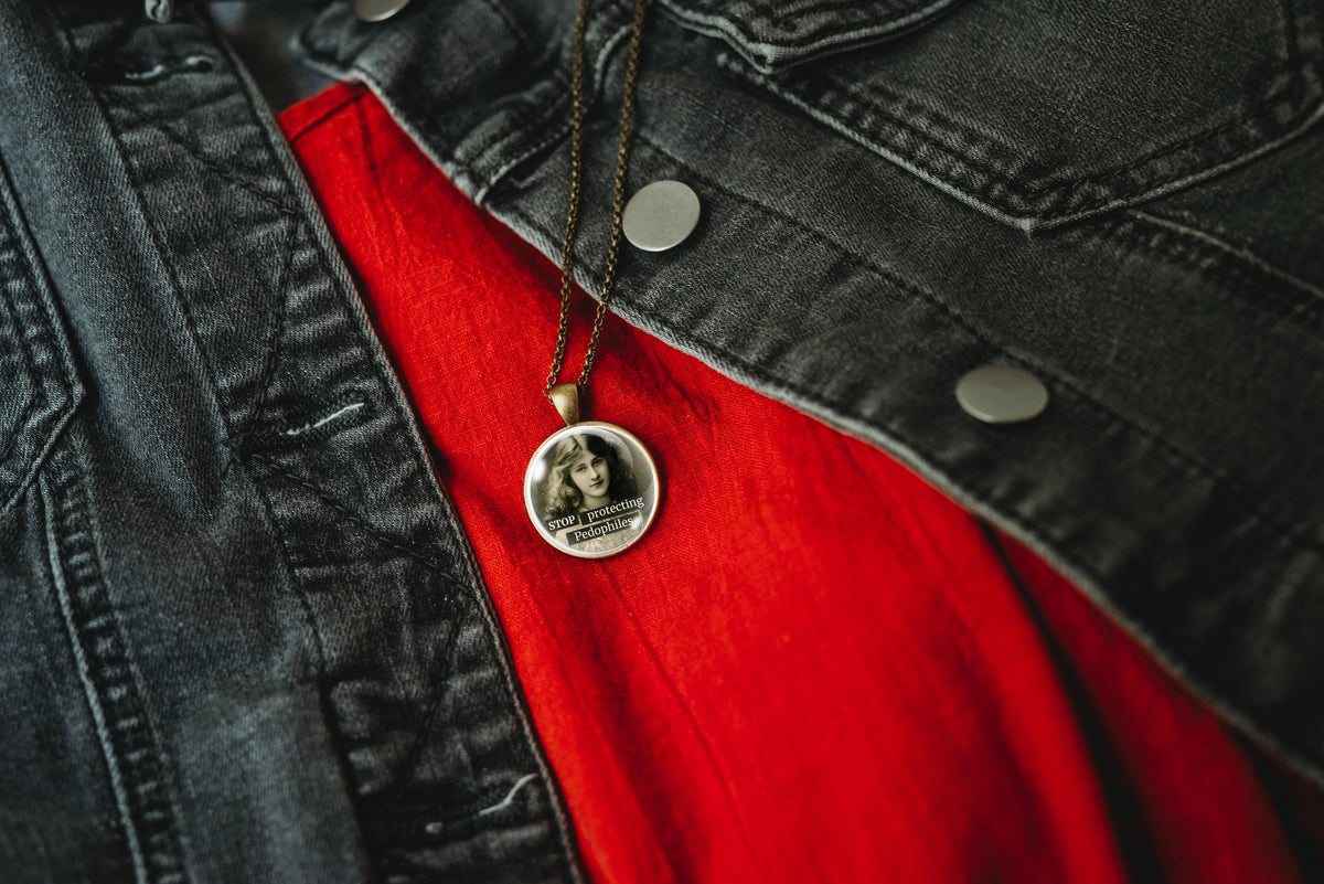 1-inch glass cabochon pendant necklace featuring “Stop Protecting Pedophiles” with a black-and-white portrait of a long-haired vintage woman on a 20-inch chain, hanging from a silver chain over a red shirt and black denim jacket.