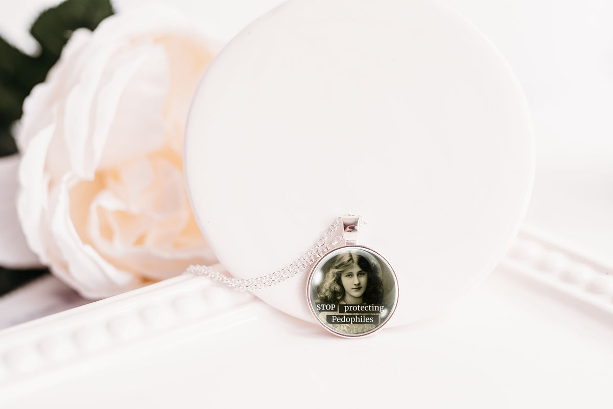 A silver-toned necklace with a round pendant displaying a black-and-white vintage portrait of a woman, resting on a white display stand next to a soft white rose.