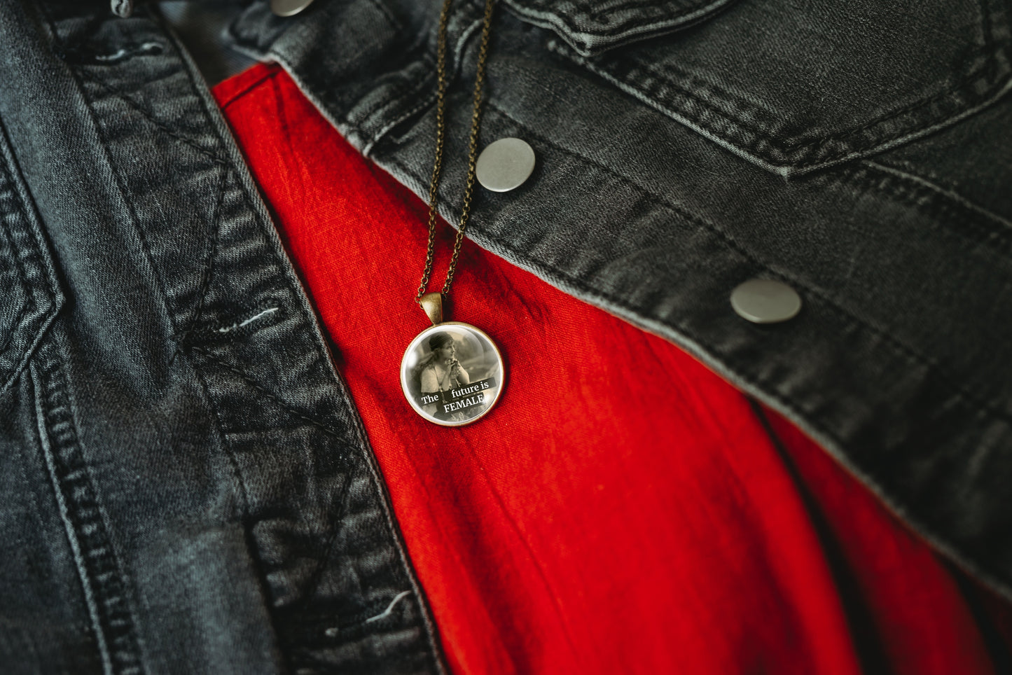 A close-up of a bronze-plated round pendant necklace on a dark chain, resting on a bright red shirt beneath an open black denim jacket.