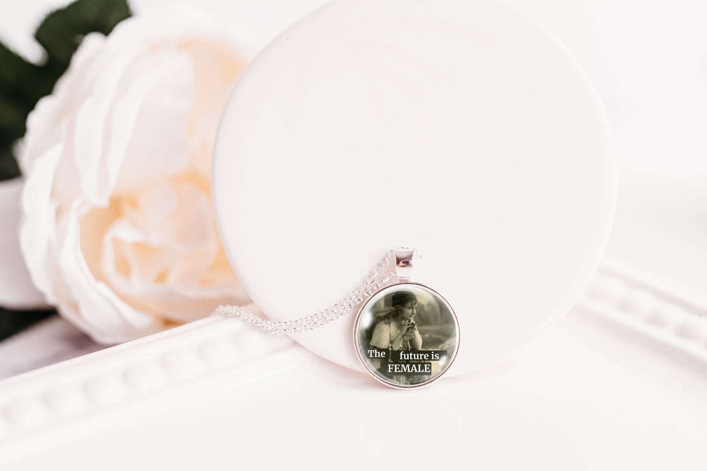 A silver-toned necklace with a round pendant resting on a white surface next to a white rose. The pendant features a printed photo of a retro boho woman and the text 'The future is FEMALE'.