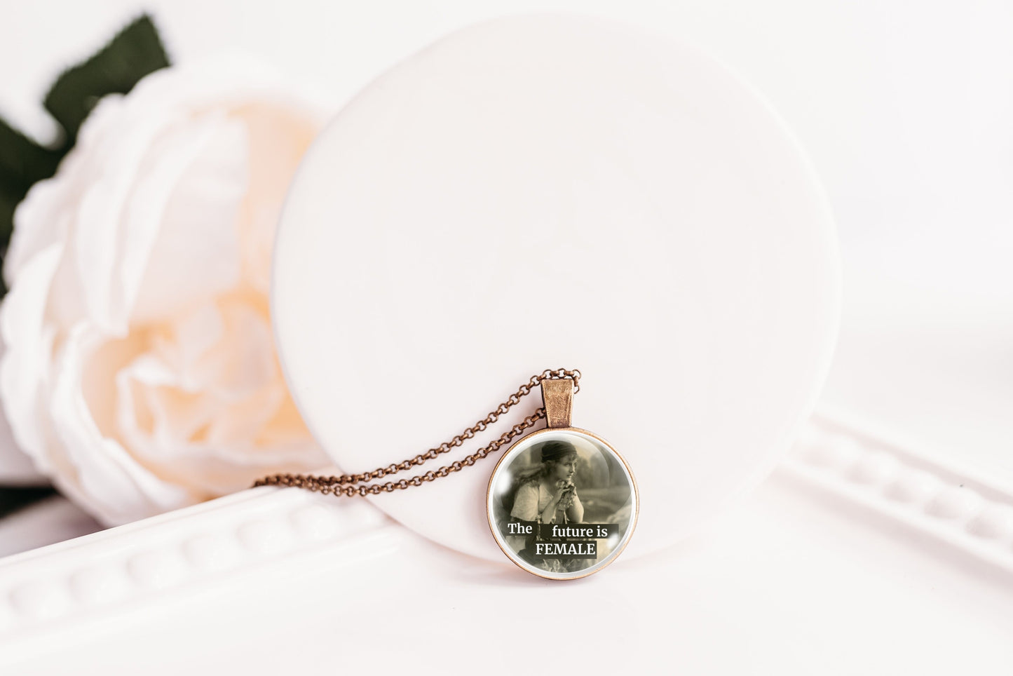 A round pendant necklace on a delicate chain, featuring a printed image of a woman and the text 'The future is FEMALE'. The pendant is displayed on a white circular stand next to a soft white rose.