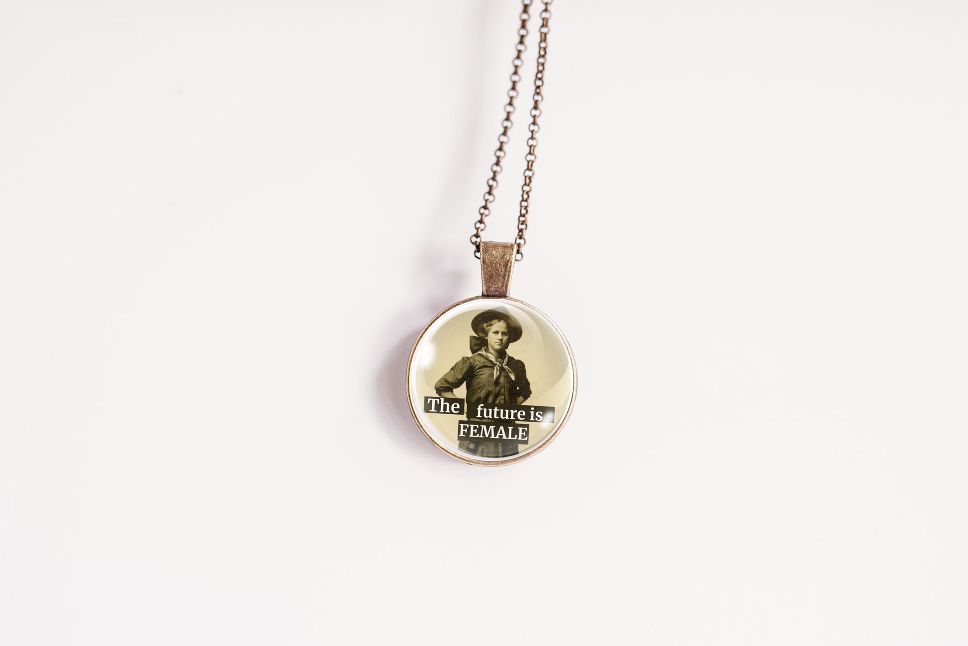A round pendant necklace on a delicate chain, featuring a vintage-style sepia portrait of a woman with the text 'the future is FEMALE' printed on the front, set against a plain white background.