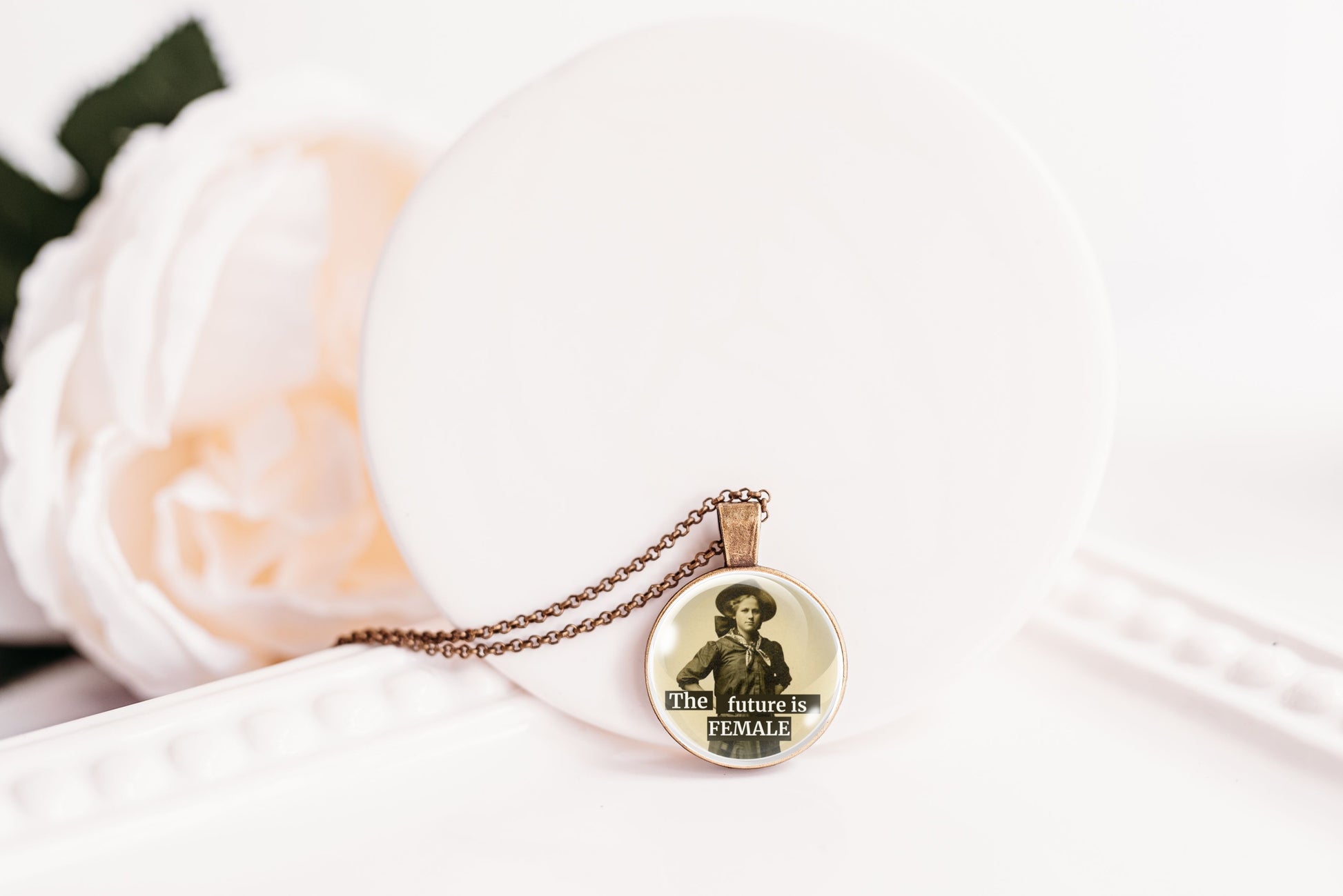 A round pendant necklace on a delicate chain, featuring a vintage-style illustration of a woman in a hat with the text 'THE future is FEMALE'. The pendant is displayed on a white circular stand next to a soft white rose.