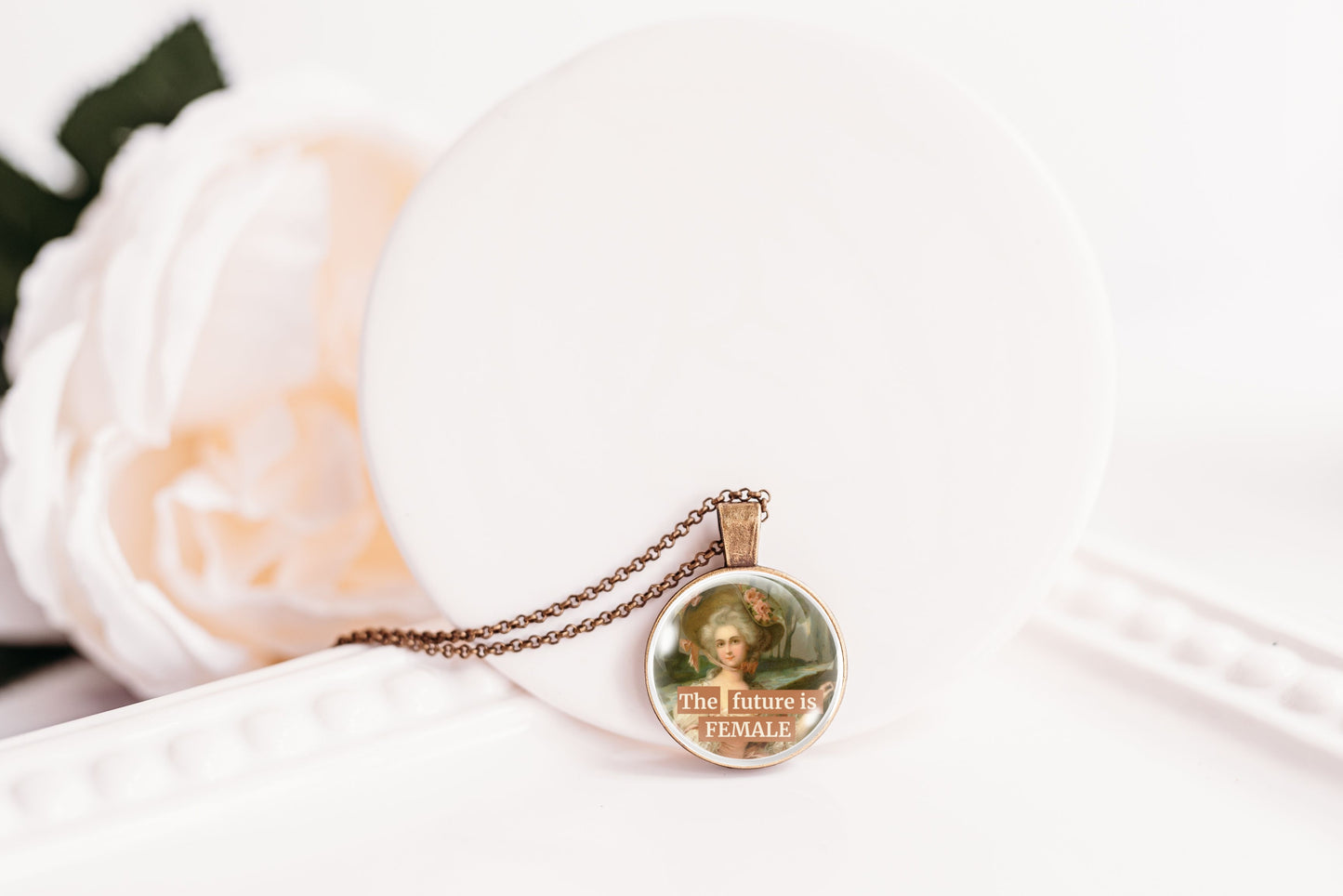 A round pendant necklace with a vintage-style portrait of a woman and the text 'The future is FEMALE' resting on a white display stand next to a soft white rose.