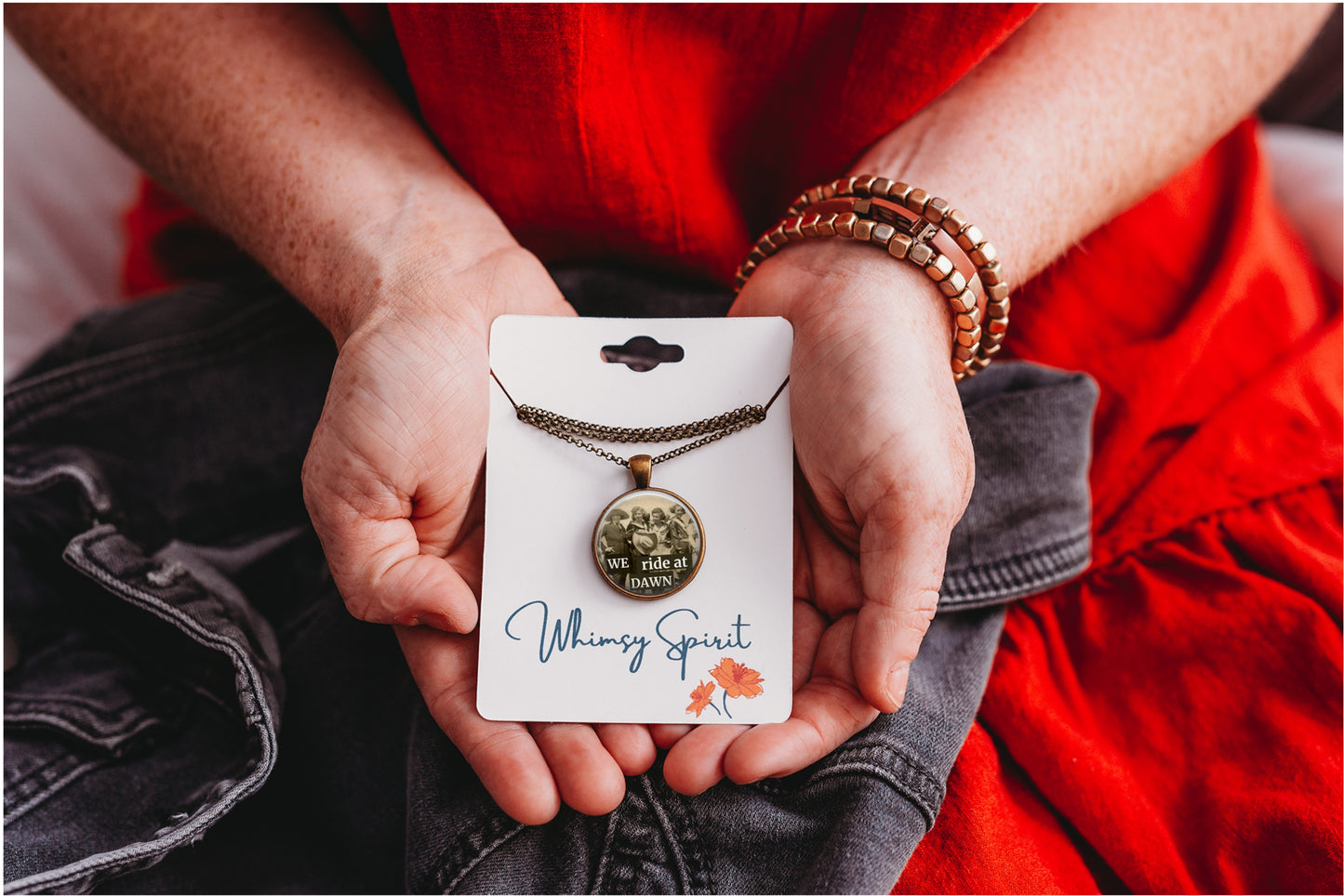 A person holding a photo pendant necklace on a white display card. The necklace has a bronze-toned chain and a round pendant featuring a black-and-white photo of four women, with the card printed with the name 'Whimsy Spirit' and a small orange flower.