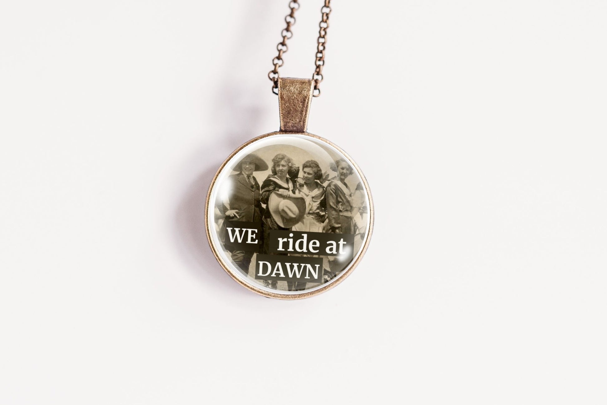 A round pendant necklace on a delicate chain, featuring a vintage-style image of a group of cowgirls with the text 'WE ride at DAWN' printed on the front.