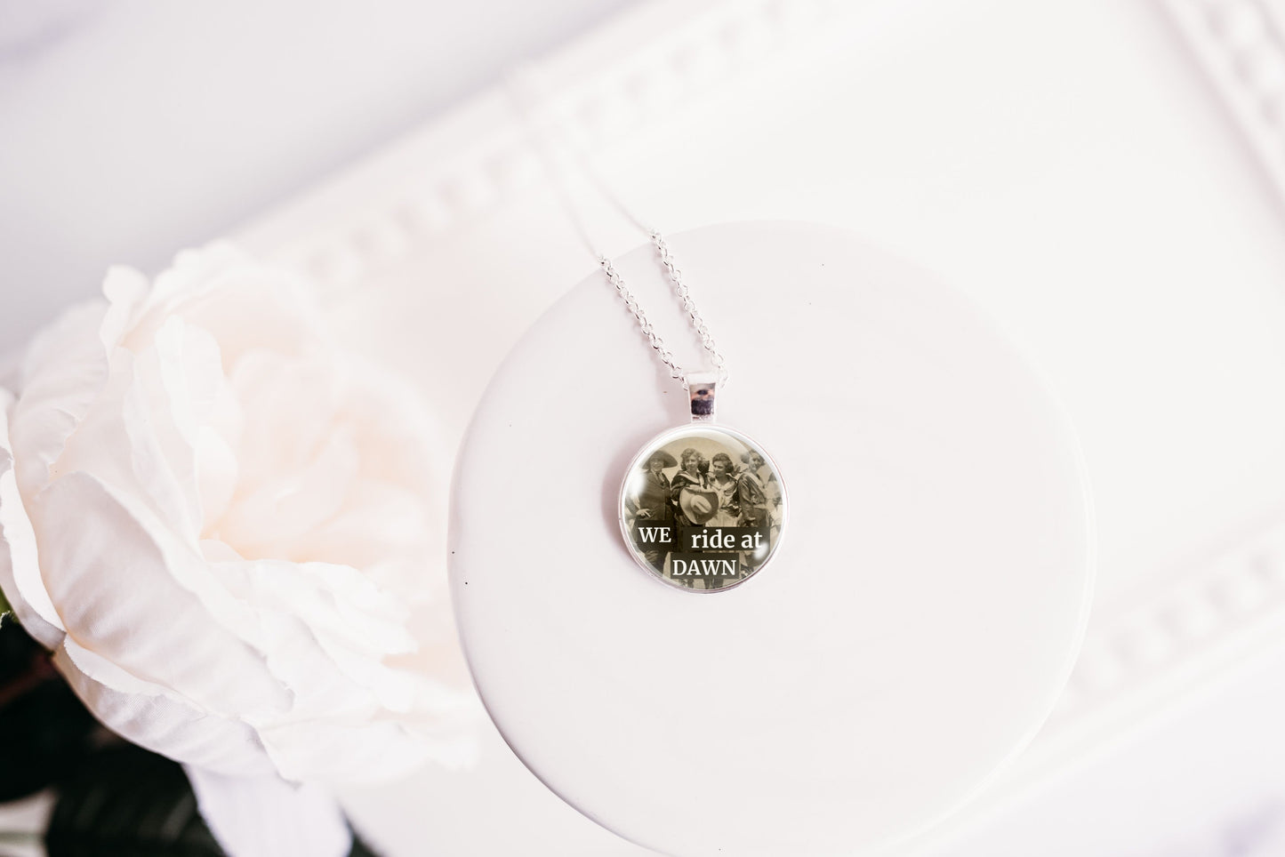 A silver necklace with a round photo pendant displayed on a white circular stand next to a white rose.