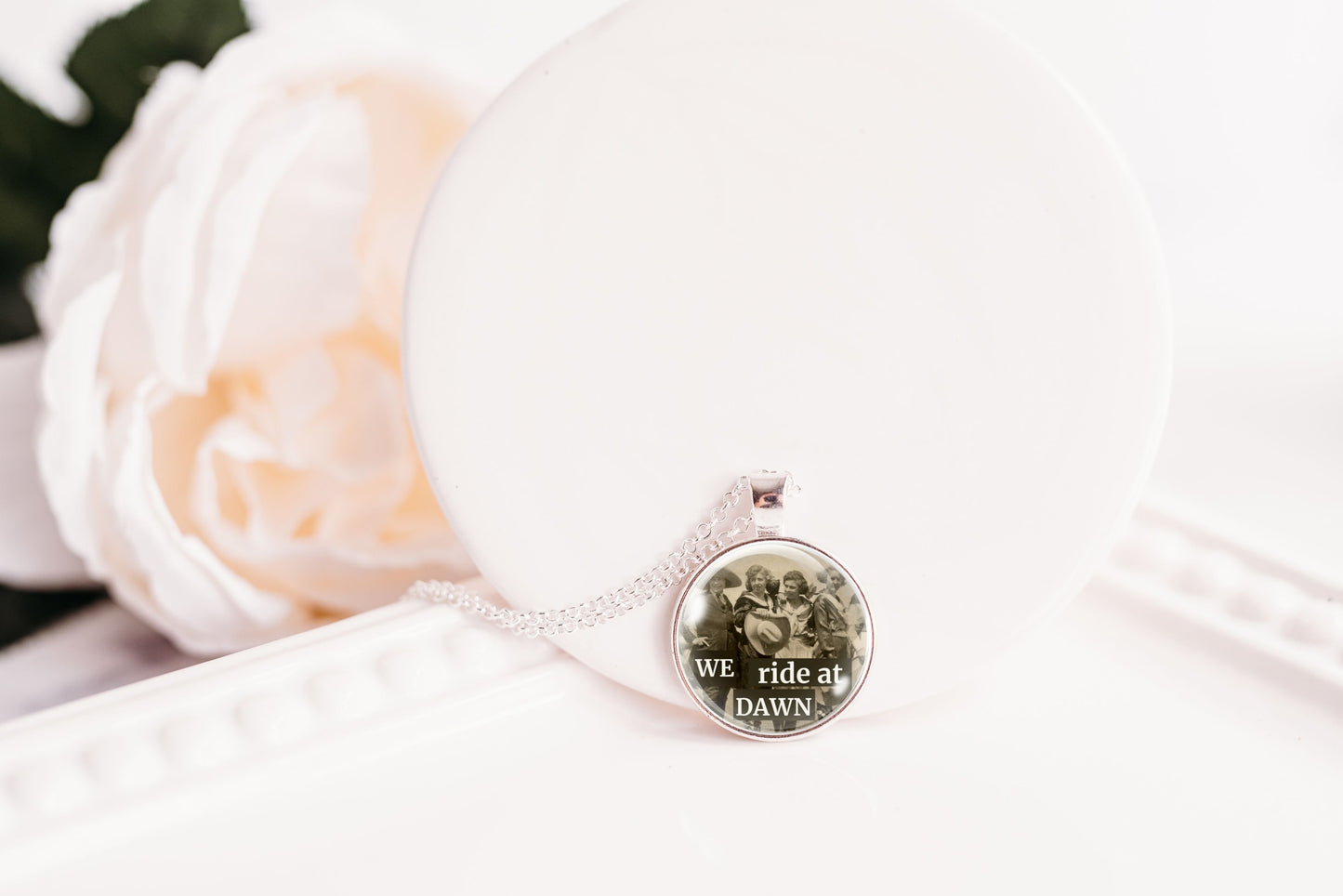 A silver-toned necklace with a round pendant resting on a white surface next to a white rose. The pendant features a printed image of four cowgirls and the text 'WE ride at DAWN'.