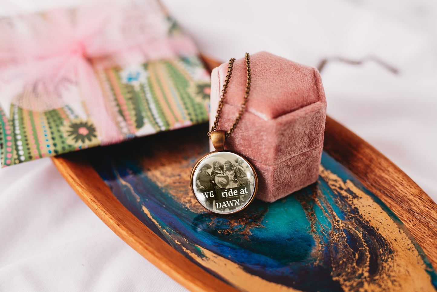 A round pendant necklace with a black-and-white image of a group of cowgirls, displayed on a pink velvet ring box resting on a decorative wooden tray with blue and gold marbled detailing.