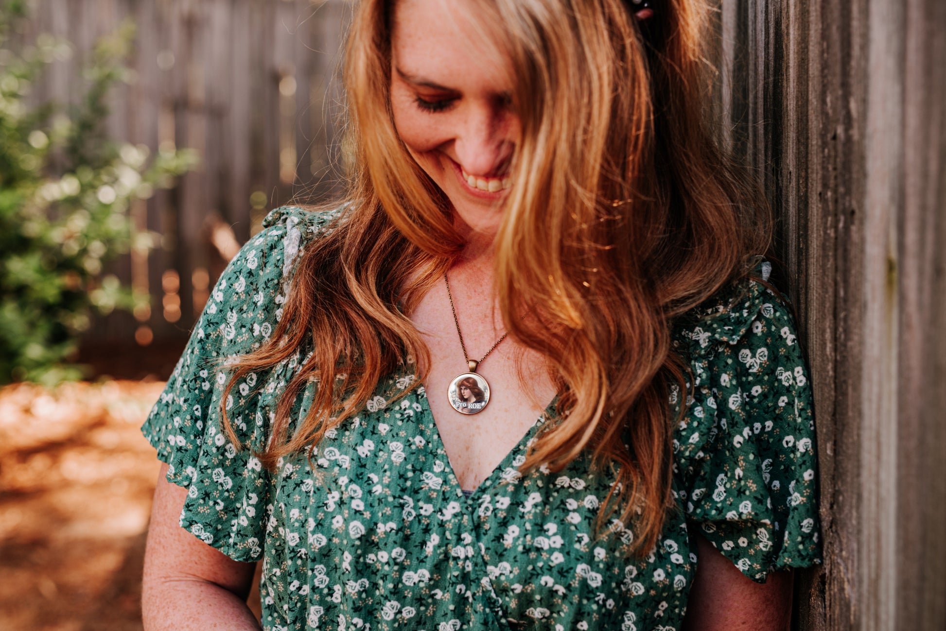A woman with long wavy light brown hair leans against a weathered wooden fence, wearing a green short-sleeve V-neck dress with a white floral pattern and a silver pendant necklace.