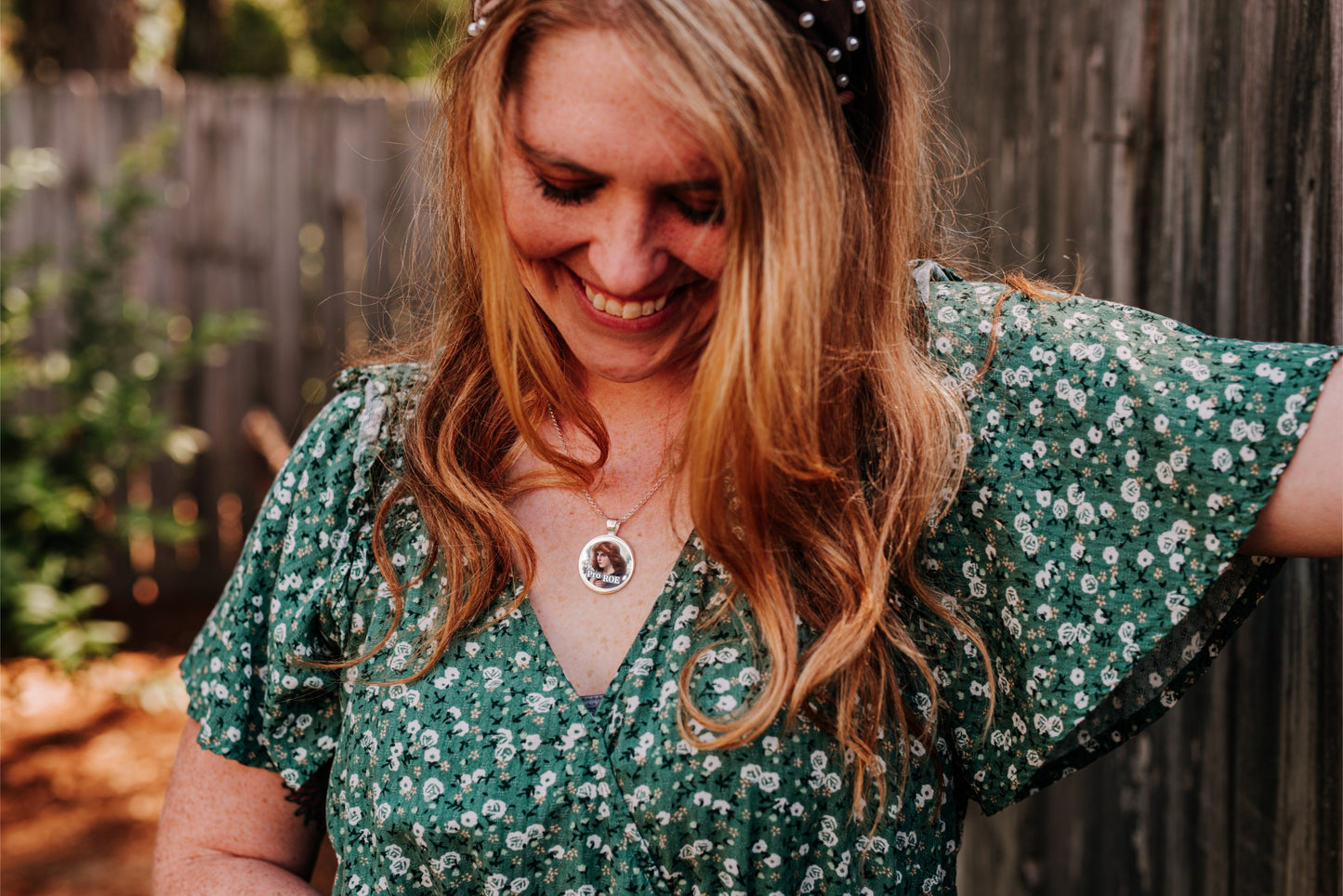 A woman with long red hair wearing a green and white floral short-sleeve dress, smiling and looking down, set against a wooden fence background.